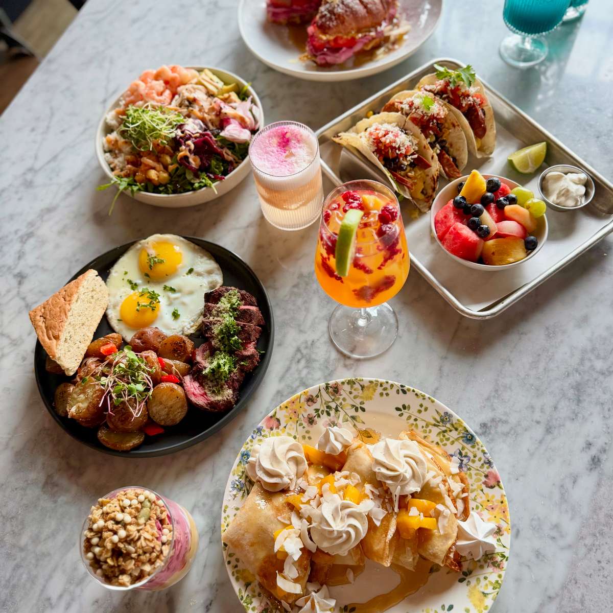colorful brunch spread with breakfast tacos, sunny-side eggs, steak and roasted potatoes, fresh fruit, pastries, and vibrant cocktails on a marble table