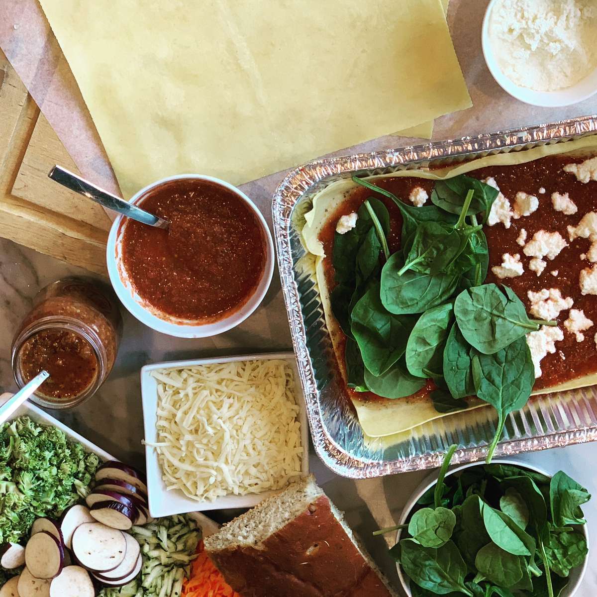 spinach lasagna prep with layers of pasta, marinara, ricotta, and fresh greens surrounded by ingredients on a kitchen prep table