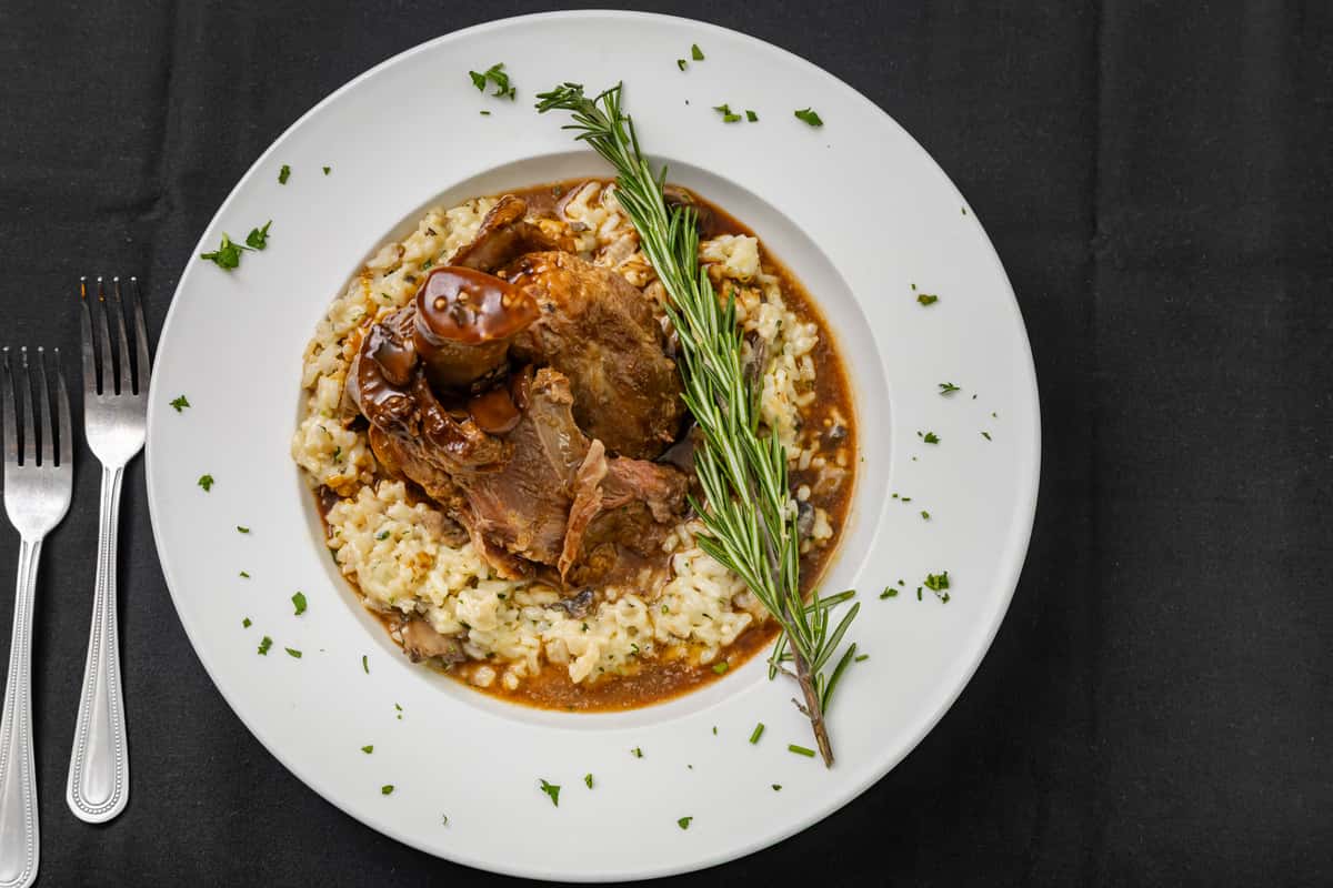 Braised lamb shank served over a bed of creamy mushroom risotto, garnished with a sprig of fresh rosemary and chopped parsley.