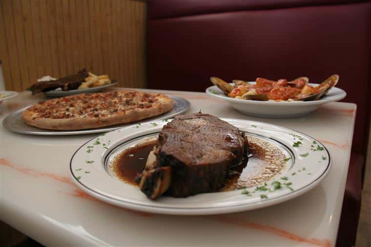 steak on a plate with a whole pizza and pasta dish in the background