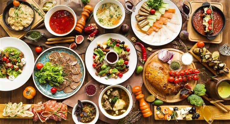 four plates with meat, vegetables, bread and cheese. four bowls of soup a wooden platewith a loaf of bread, onions and spices. spices scattered in between dishes