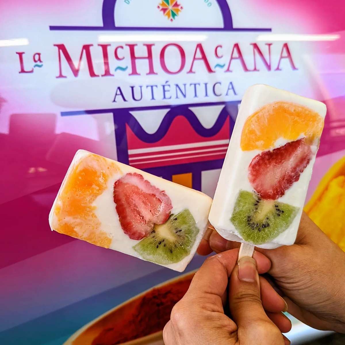 Two fruit-flavored popsicles with strawberries, kiwi, and orange slices, held up in front of the "La Michoacana Auténtica" logo.