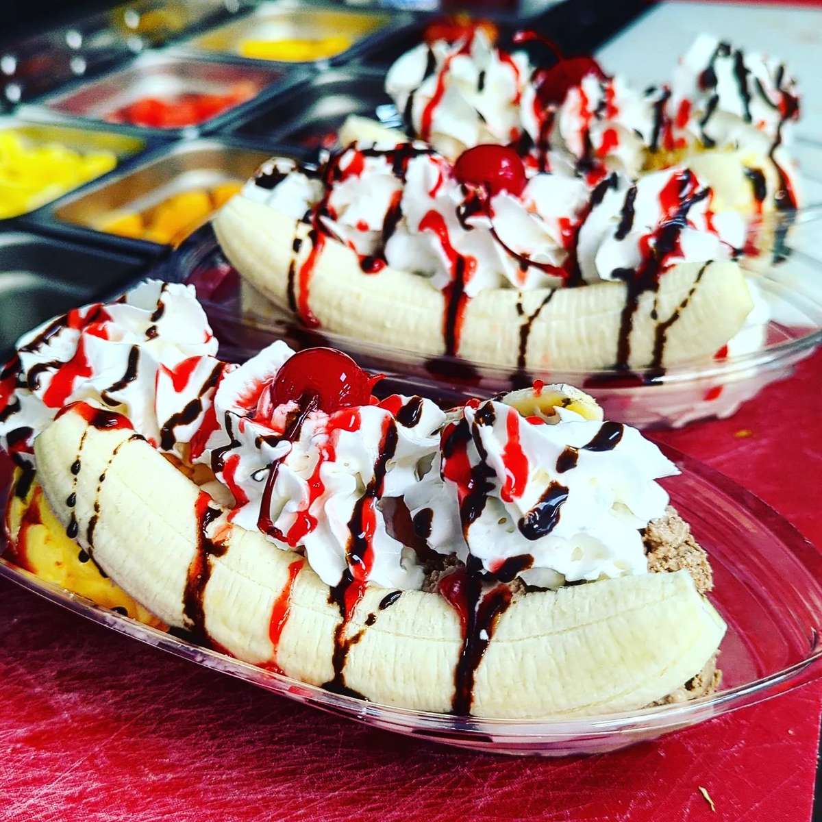 Two banana split sundaes topped with whipped cream, chocolate and strawberry syrup, and cherries in clear plastic dishes.