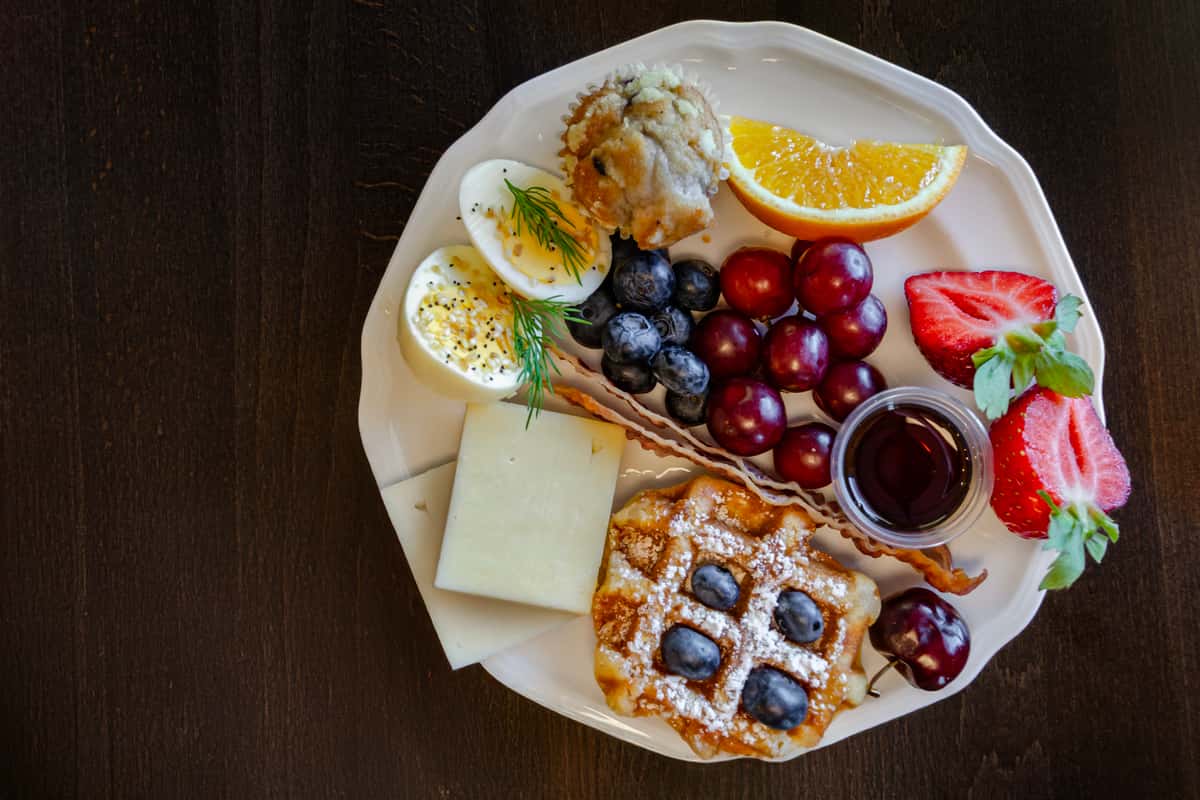 Overhead view of a brunch plate with a mini waffle, hard-boiled eggs, cheese, bacon, grapes, blueberries, strawberries, a muffin, and an orange wedge — ideal for full-service brunch menus or bed-and-breakfast offerings.