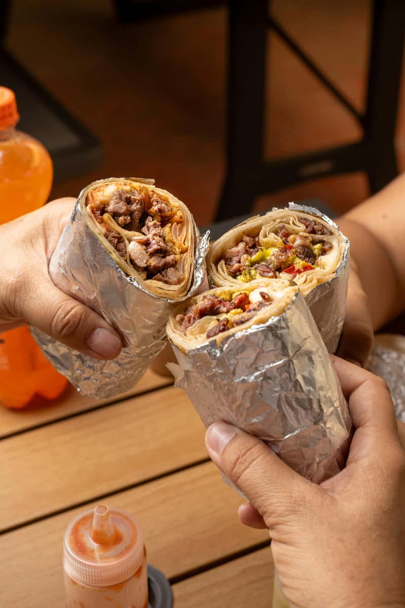 three hands holding and clinking foil-wrapped beef burritos over a wooden table