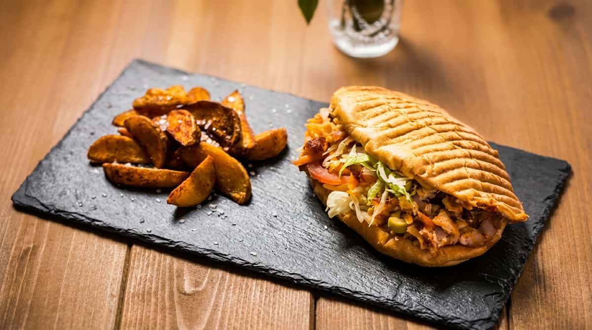 Sandwich with meat, lettuce, tomatoes, and red cabbage, served with potato wedges on a slate plate.