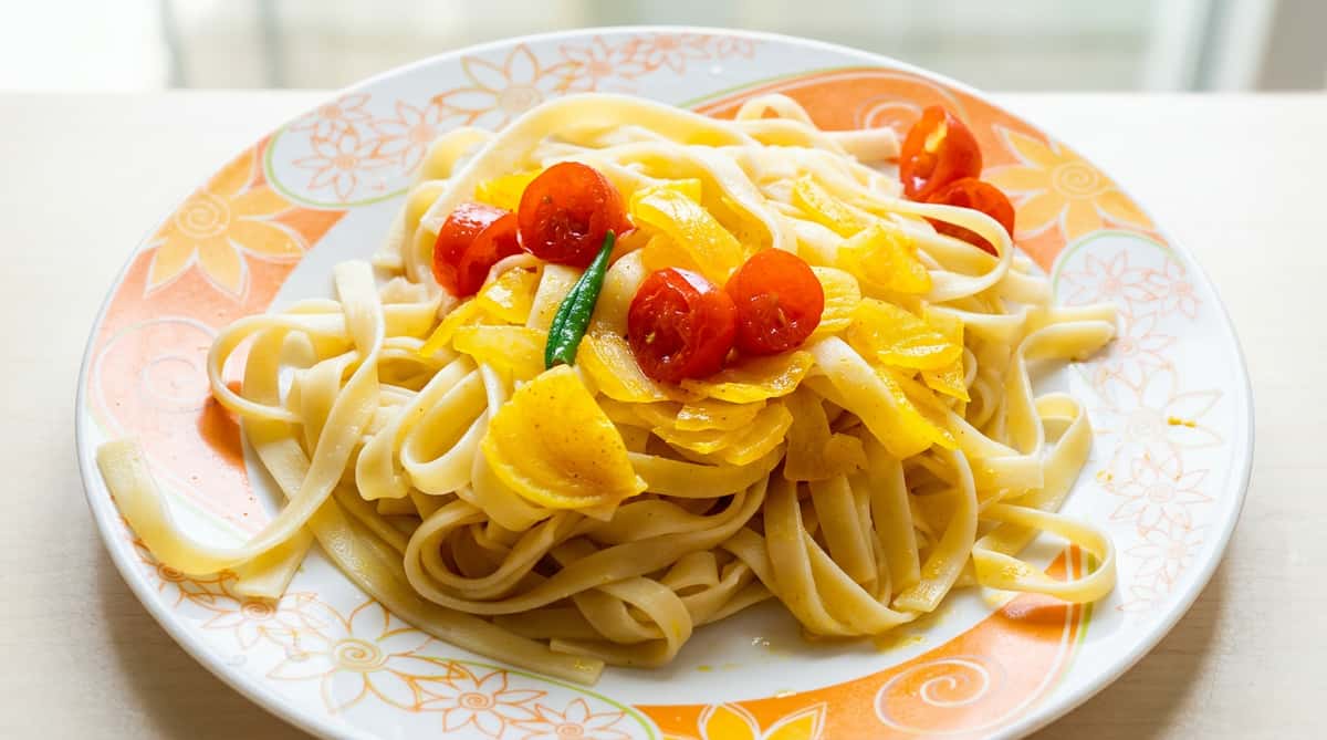 Fettuccine pasta with sautéed yellow bell peppers, cherry tomatoes, and a green chili on a floral plate.