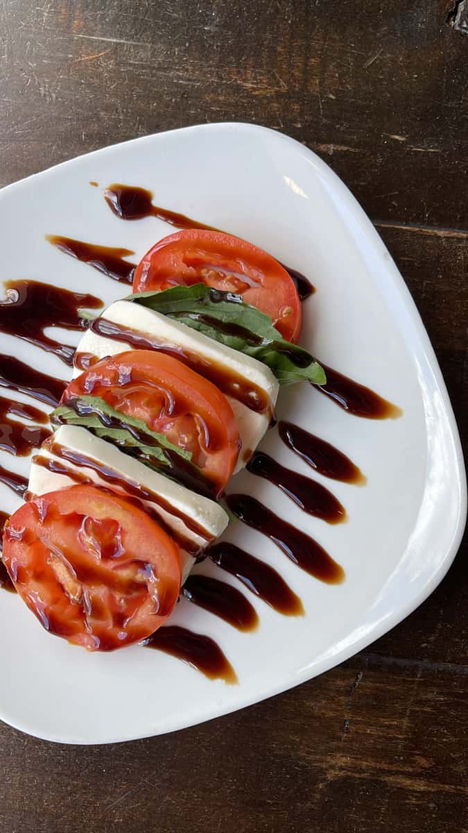 A classic Caprese salad featuring layers of ripe tomatoes, fresh mozzarella, and basil leaves, elegantly arranged on a white plate and finished with a rich balsamic glaze.