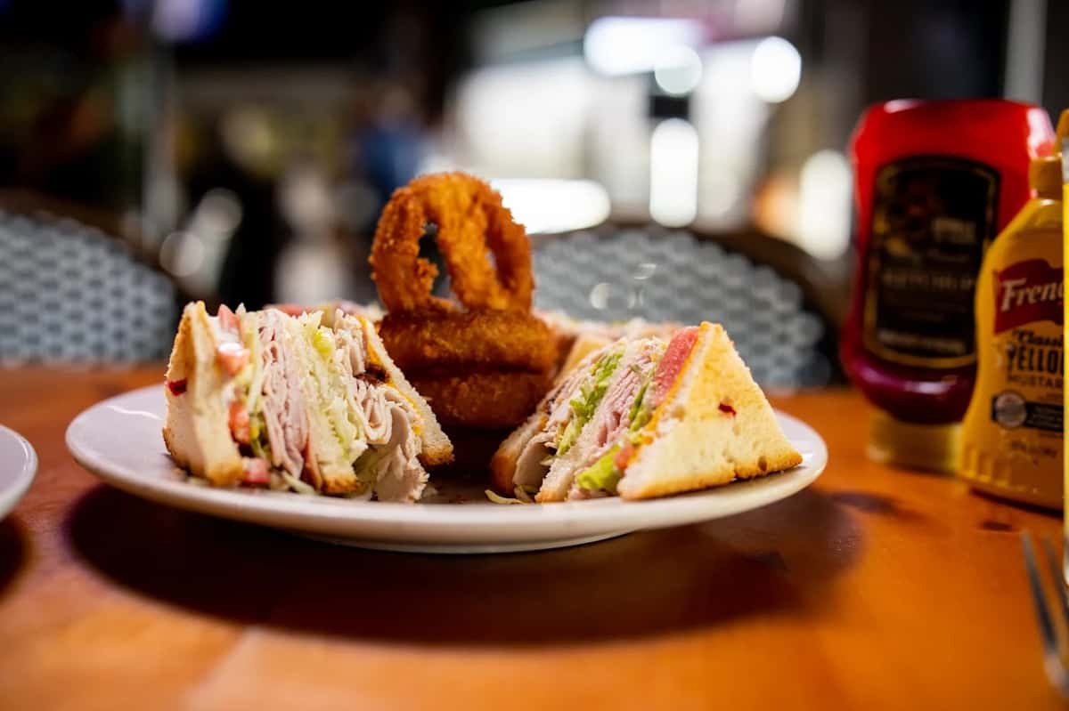 Club sandwich stacked with turkey, lettuce, tomato, and bacon, served with crispy onion rings on a white plate.