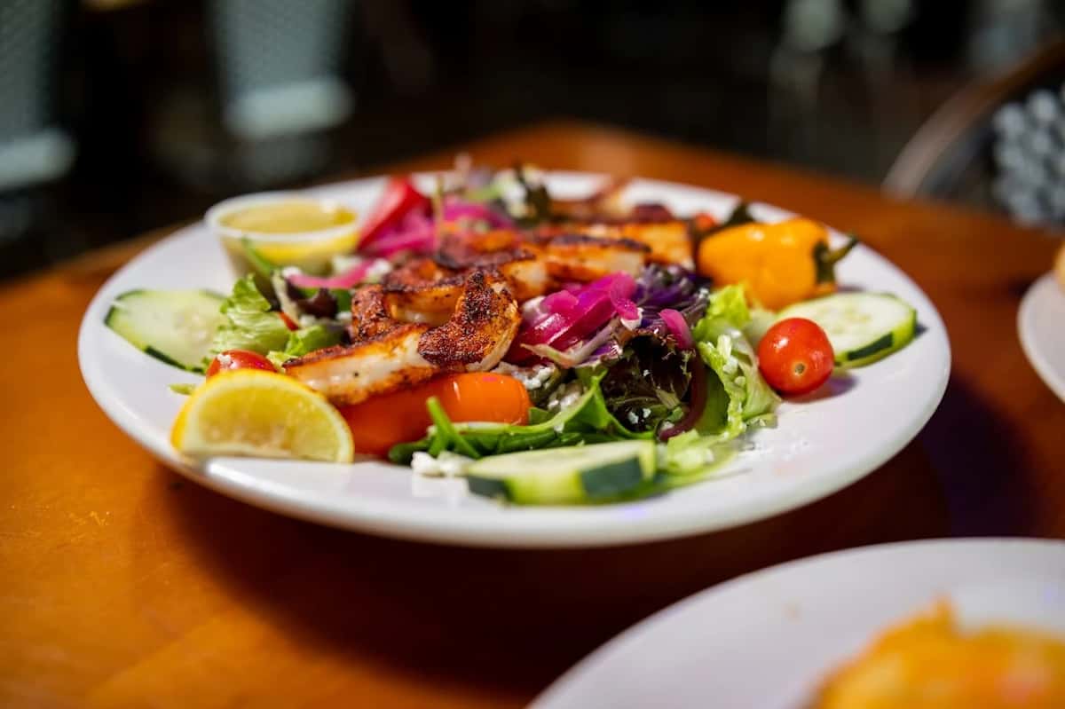 Grilled shrimp salad with mixed greens, cherry tomatoes, cucumber, red onion, and lemon wedges served with a side of dressing.