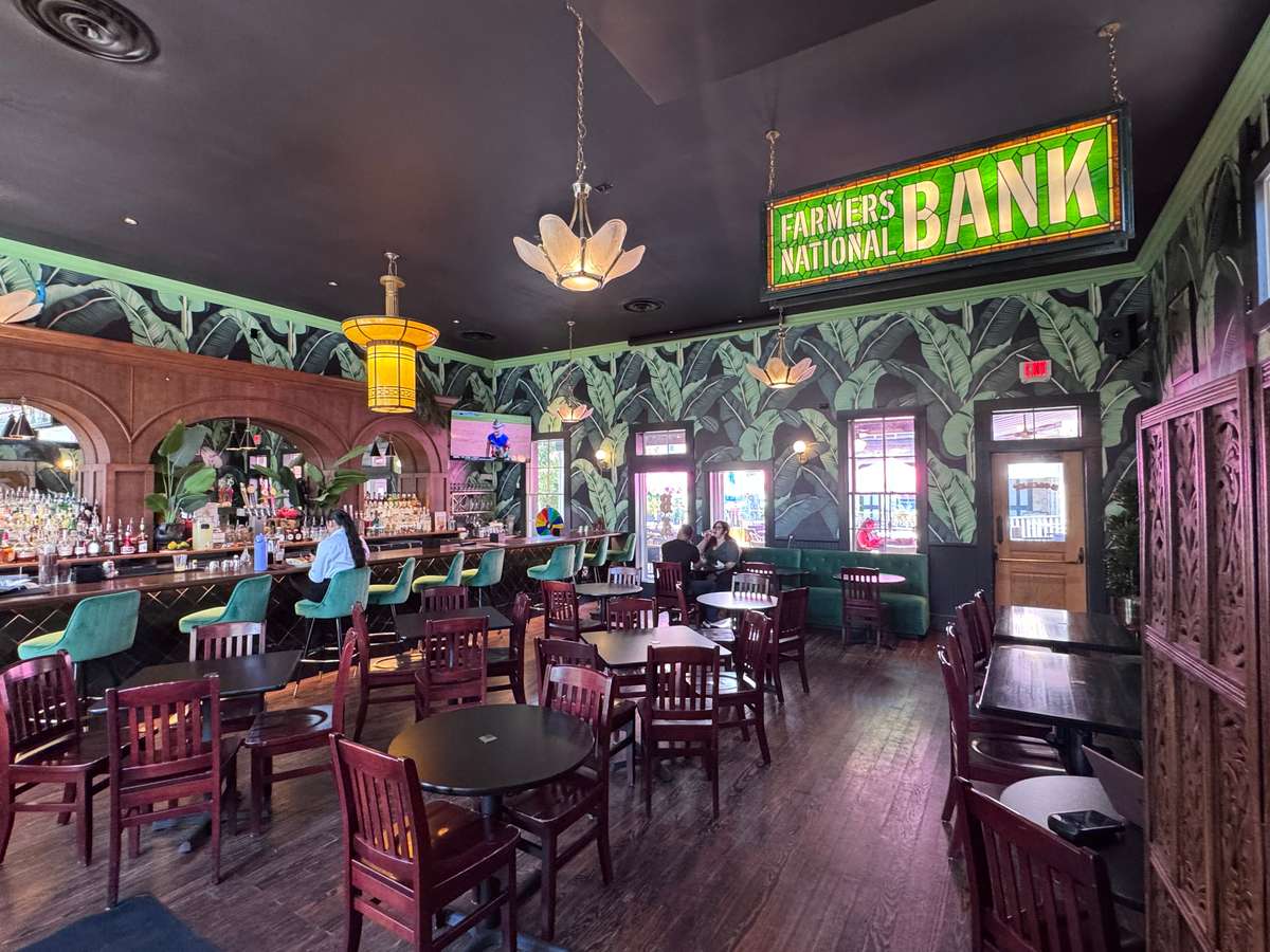 Vibrant Bombay Bicycle Club interior featuring tropical green leaf wallpaper, emerald bar stools, dark wood tables, and a vintage "FARMERS NATIONAL BANK" sign.