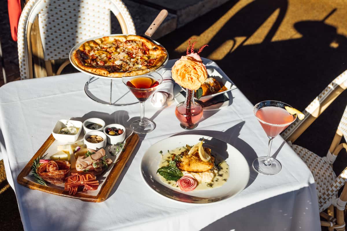 Outdoor table set with pizza, charcuterie board, fish entrée, and pink cocktails in bright sunlight.
