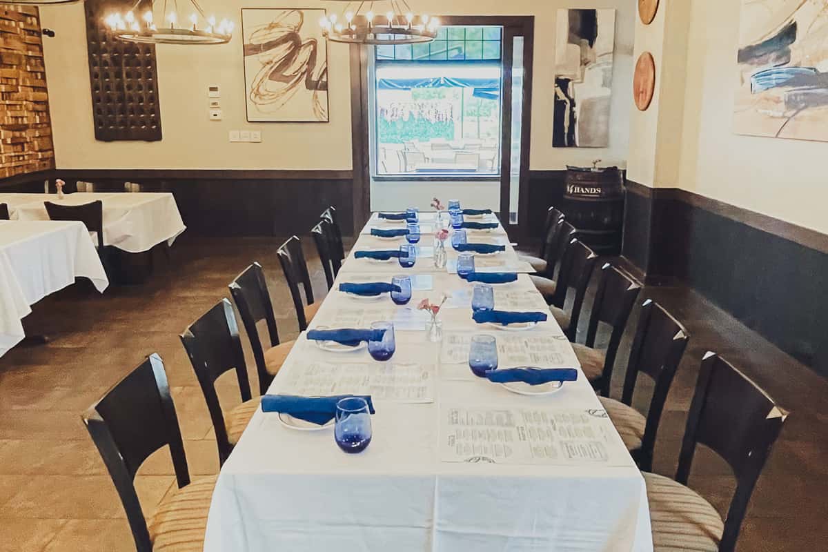 Long table set for private dining in the Barrel Room at Osteria Italian Seafood in Louisville. Features navy napkins, blue glassware, and soft chandelier lighting with a view to the patio.