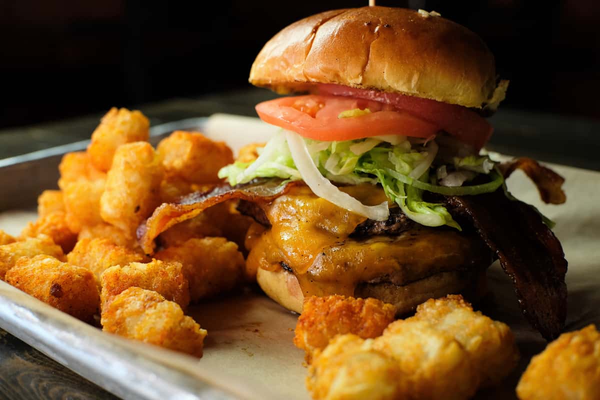 Cheeseburger topped with lettuce, tomato, onions, and bacon, served with golden tater tots on a metal tray.