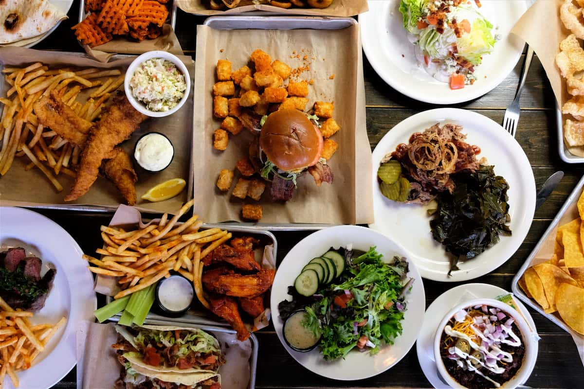 Assorted American comfort food including burgers, tacos, wings, fries, salad, and a variety of side dishes on a wooden table.