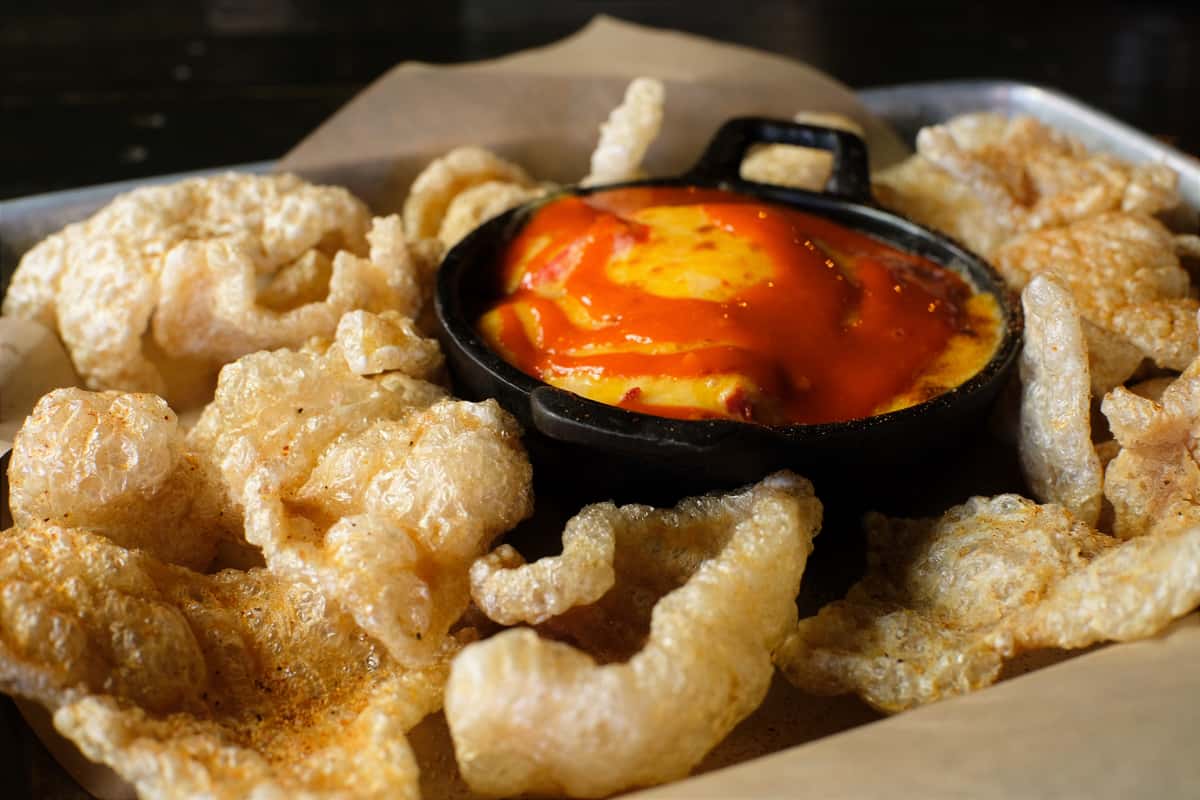 Crispy pork rinds served with a skillet of melted cheese dip topped with vibrant red hot sauce.