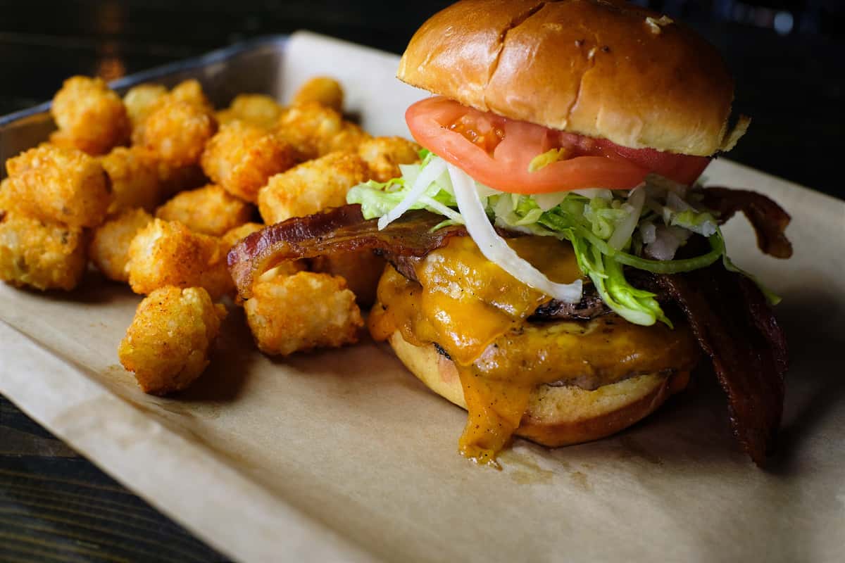 Cheeseburger topped with bacon, lettuce, tomato, and onion, served with crispy golden tater tots on a parchment-lined tray.