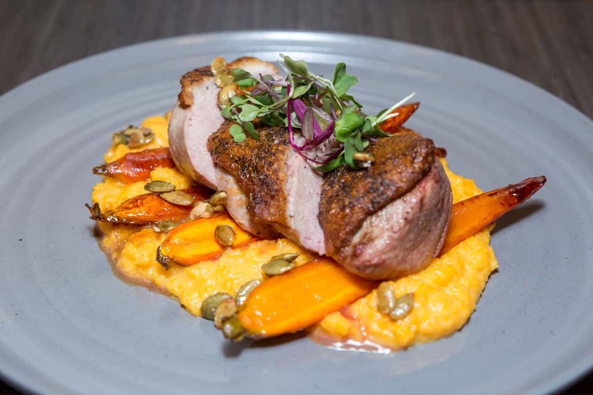 This image features a beautifully plated entrée showcasing thick slices of perfectly cooked pork tenderloin, tender and juicy with a lightly seared exterior. The meat is served over a vibrant bed of roasted vegetables—featuring carrots and possibly squash—coated in a rich, golden sauce. A garnish of fresh microgreens adds a pop of color and a touch of freshness, completing this refined and hearty dish.