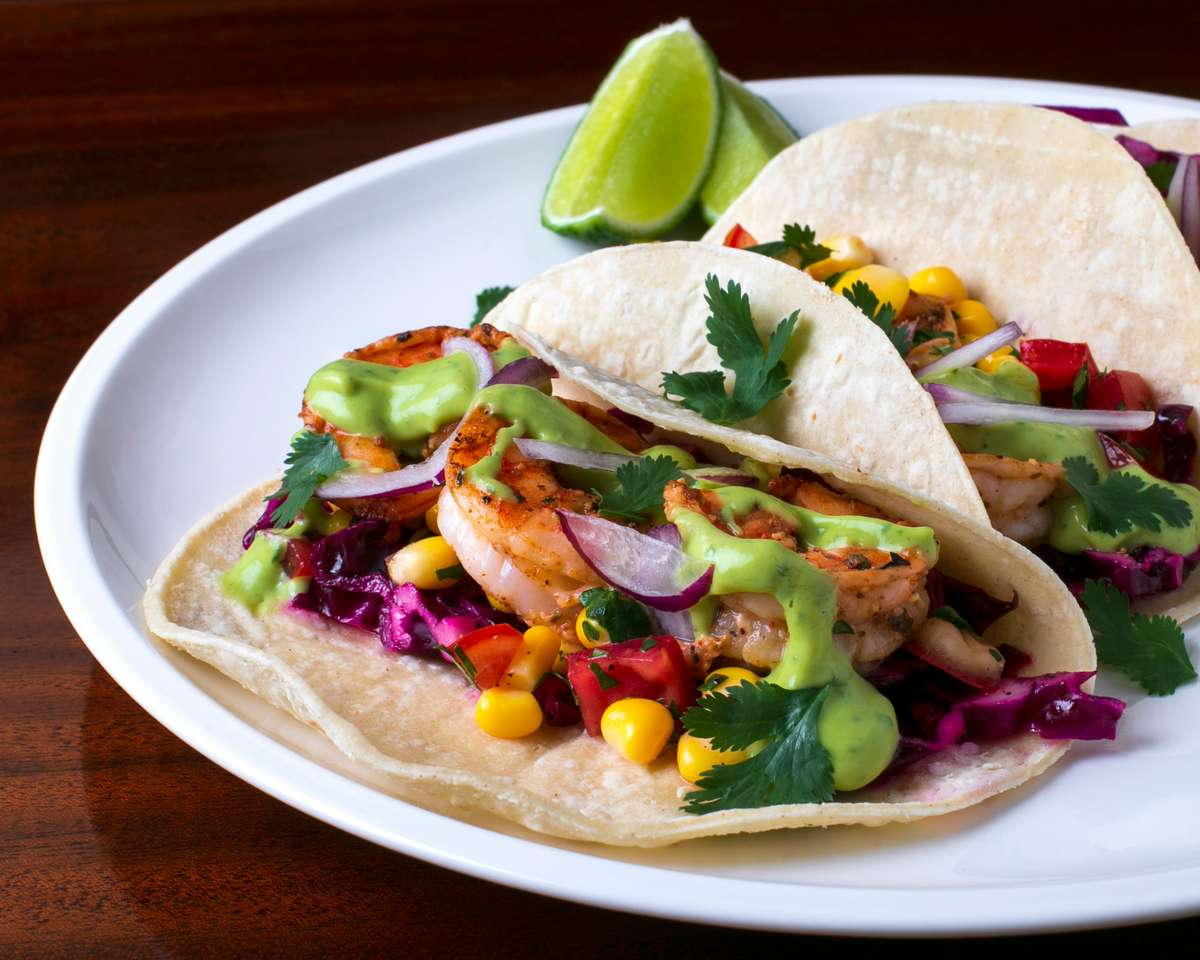 Shrimp tacos loaded with grilled prawns, shredded cabbage, corn, red onions, and avocado-lime crema, garnished with cilantro and served with fresh lime wedges.