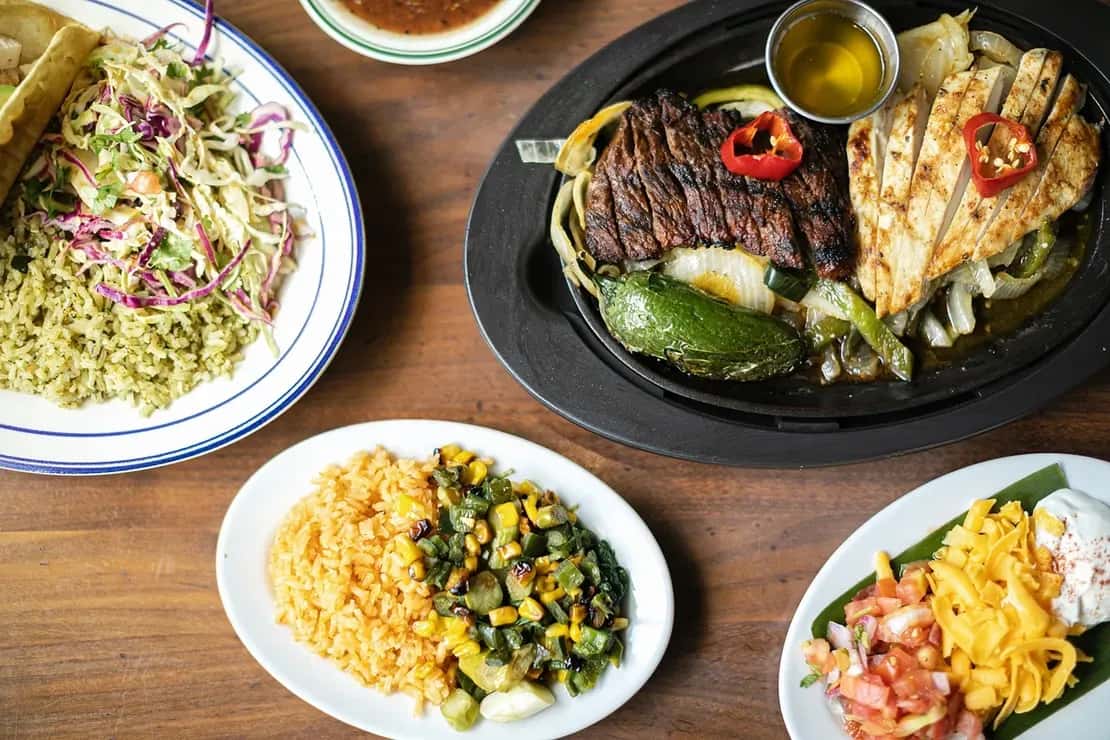 Sizzling steak and grilled chicken fajitas with sautéed peppers and onions, served alongside Mexican rice, elote-style corn, tacos, and fresh toppings on a rustic wooden table