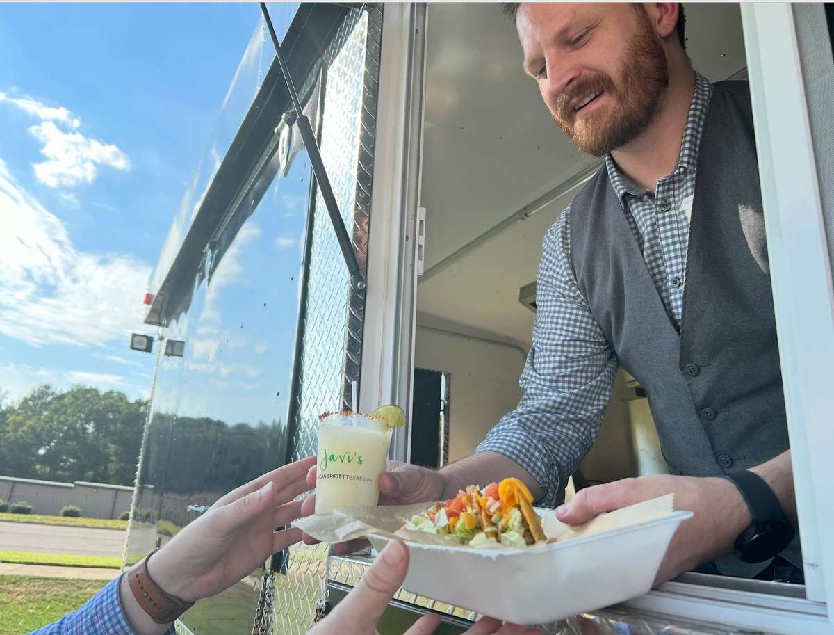 Food truck service window with staff handing a margarita and street-style tacos in a takeout container, capturing a casual outdoor dining experience with fresh Mexican cuisine.