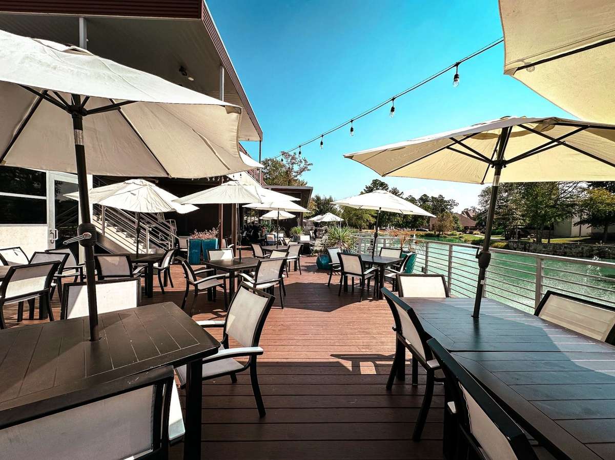 Spacious waterfront restaurant patio with outdoor seating, sun umbrellas, and string lights, featuring a wooden deck overlooking a calm canal on a bright sunny day.