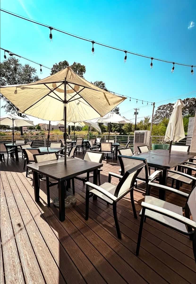 Outdoor restaurant patio with wooden deck seating, large cream umbrellas, and string lights under a clear blue sky, creating a relaxed al fresco dining atmosphere.