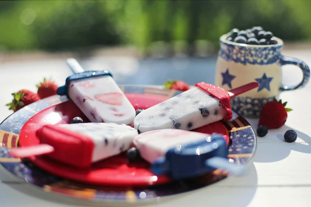 Frozen Fusion's festive red, white, and blue berry popsicles on a star-spangled plate with fresh fruit outdoors.