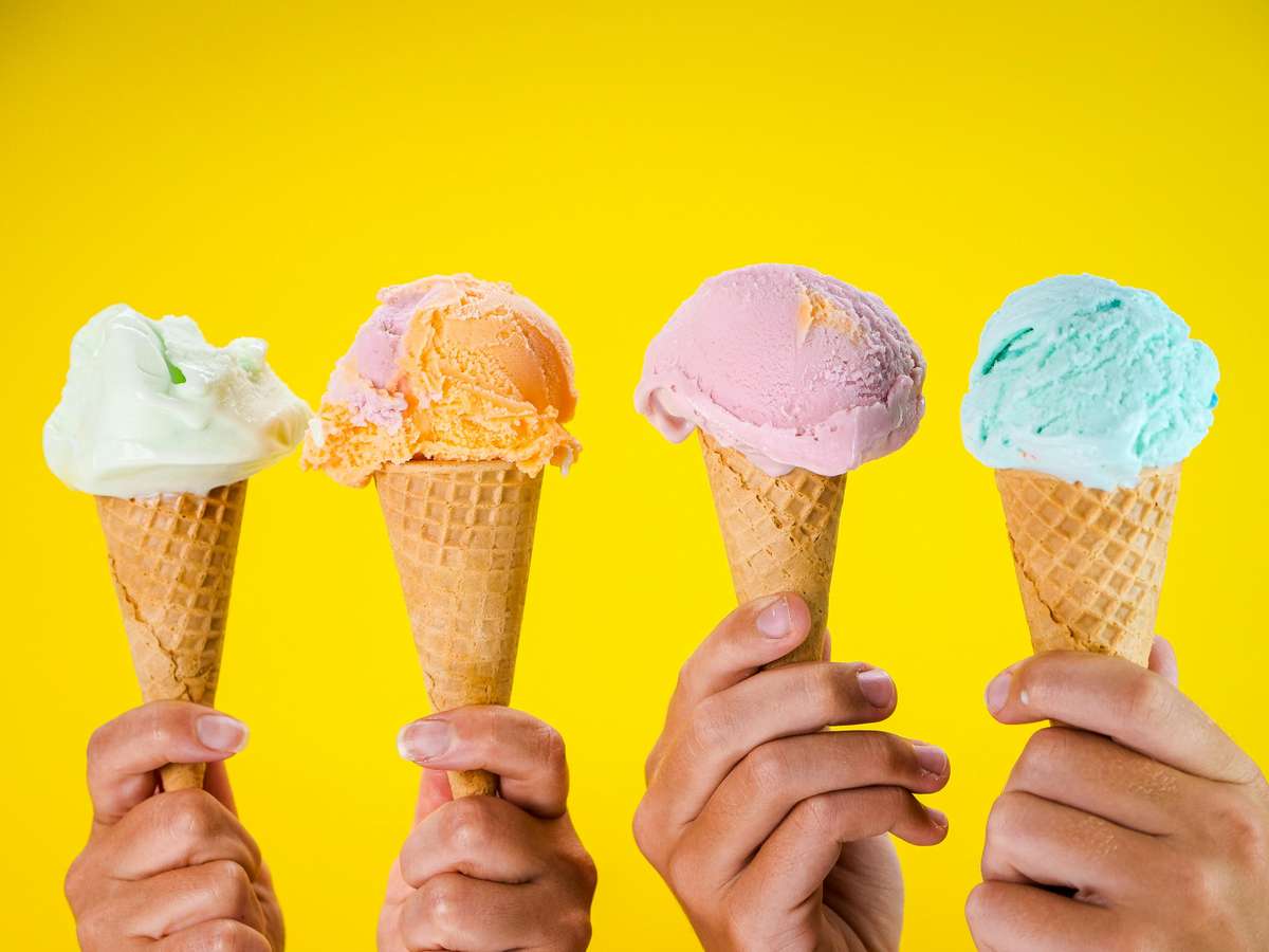 Four hands hold individual pastel-colored ice cream cones against a vivid yellow background, featuring Frozen Fusion's refreshing treats.