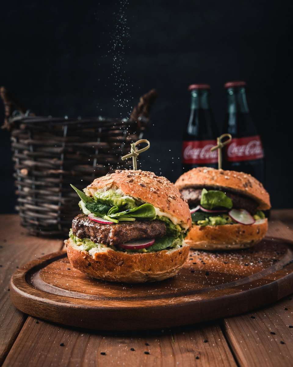 Gourmet beef burgers with avocado spread, spinach, and radish on seeded buns, served on a wooden board with Coca-Cola bottles in a rustic restaurant setting.