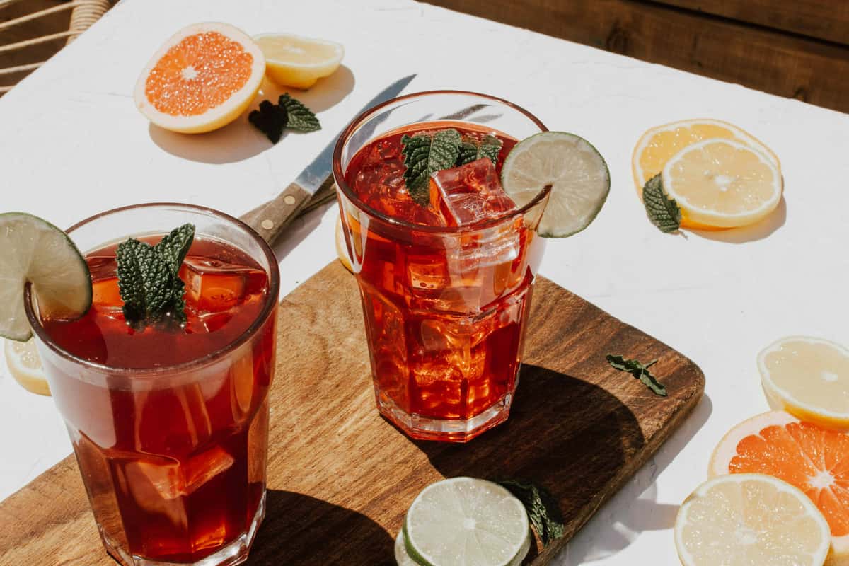 Two glasses of iced tea with lime slices, mint leaves, and ice cubes on a wooden board, surrounded by citrus slices.