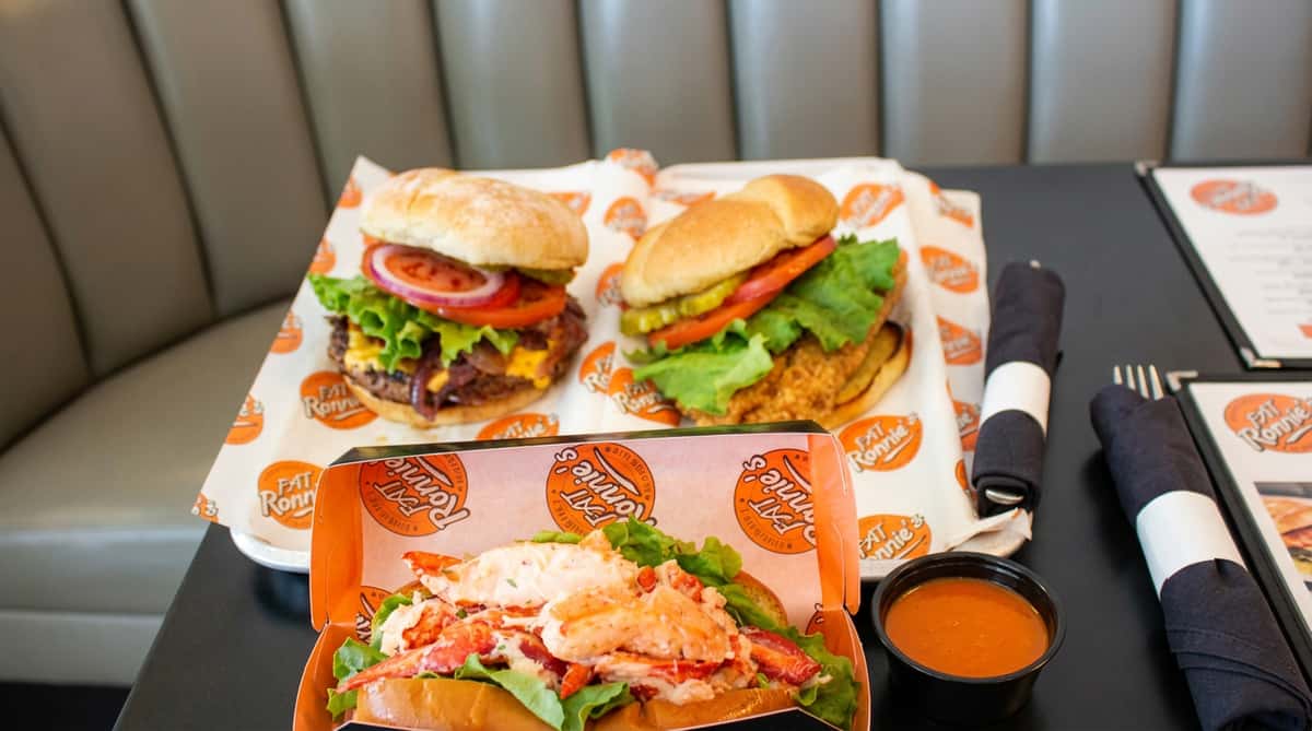 Three sandwiches on paper with "Fat Ronnie's" logo. A lobster roll, cheeseburger, and fried chicken sandwich with a side of sauce.