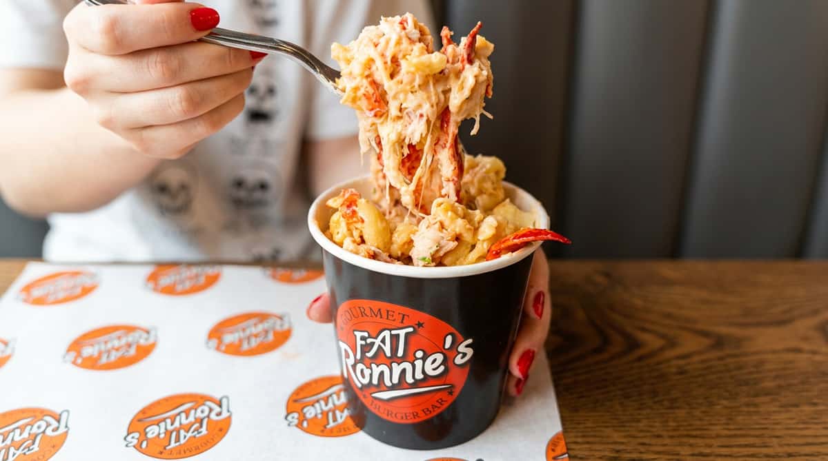 Lobster mac and cheese in a black cup from "Fat Ronnie's Burger Bar," with a fork lifting a creamy bite.