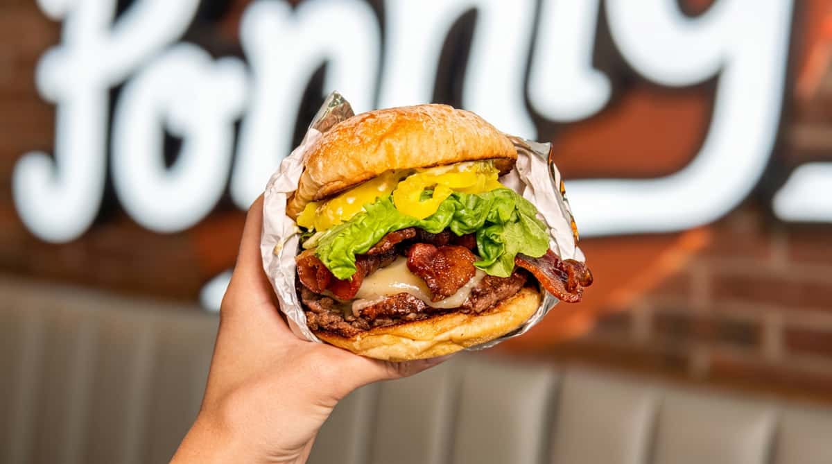 Hand holding a burger with two patties, bacon, melted cheese, lettuce, and banana peppers wrapped in foil.