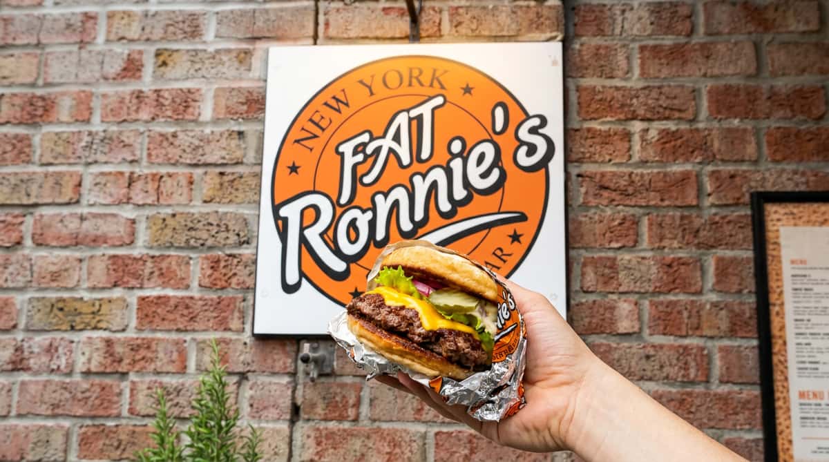 Hand holding a cheeseburger with lettuce, pickles, and onion wrapped in foil, against Fat Ronnie's Burger Bar sign.