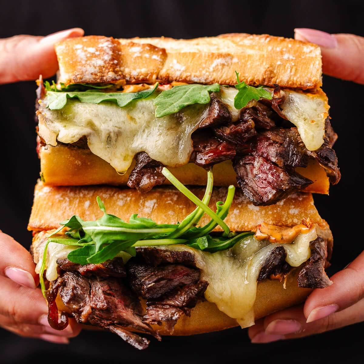 Close-up of stacked steak sandwich with melted cheese and arugula on toasted bread, held in both hands.