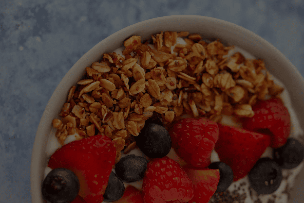 Bowl of creamy yogurt topped with granola, fresh strawberries, raspberries, and blueberries on blue surface