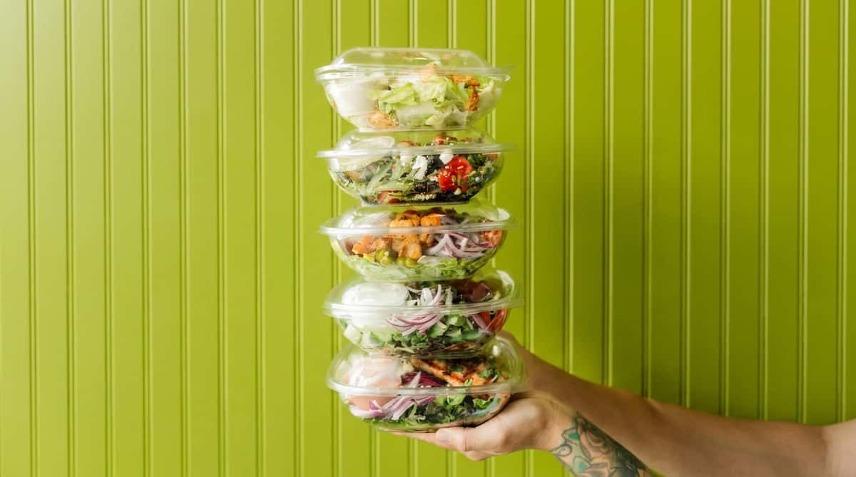 Hand holding a stack of five clear plastic containers with various colorful salads against a green wall.