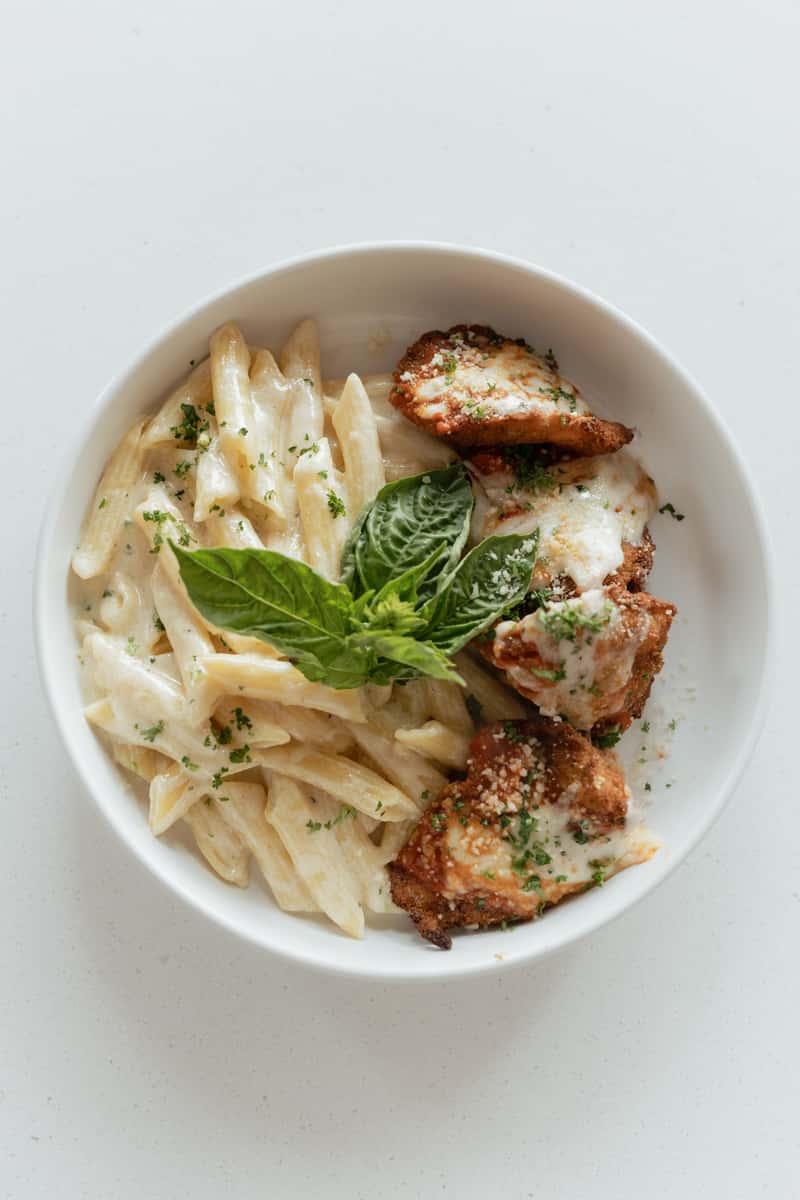 A bowl of creamy penne pasta (likely Alfredo) served with pieces of breaded chicken topped with melted cheese and herbs, garnished with fresh basil.
