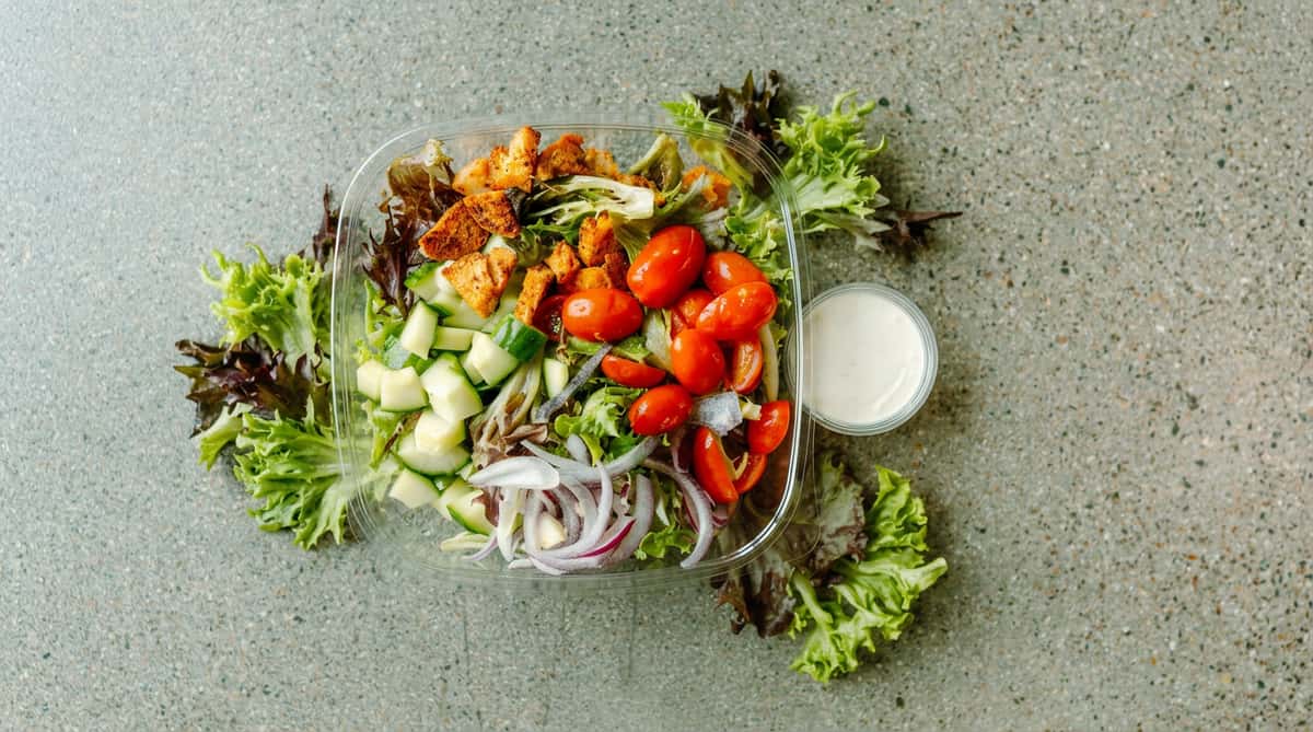 Salad in a clear plastic container with lettuce, cucumbers, cherry tomatoes, croutons, and red onions. A small dressing cup is on the side.