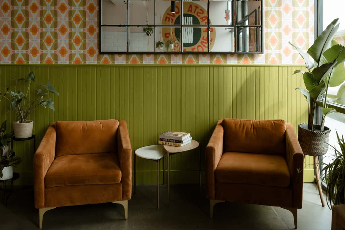 A cozy seating area in a modern café, featuring two brown upholstered chairs, a small side table with books, green paneled walls, patterned wallpaper, and several indoor plants.