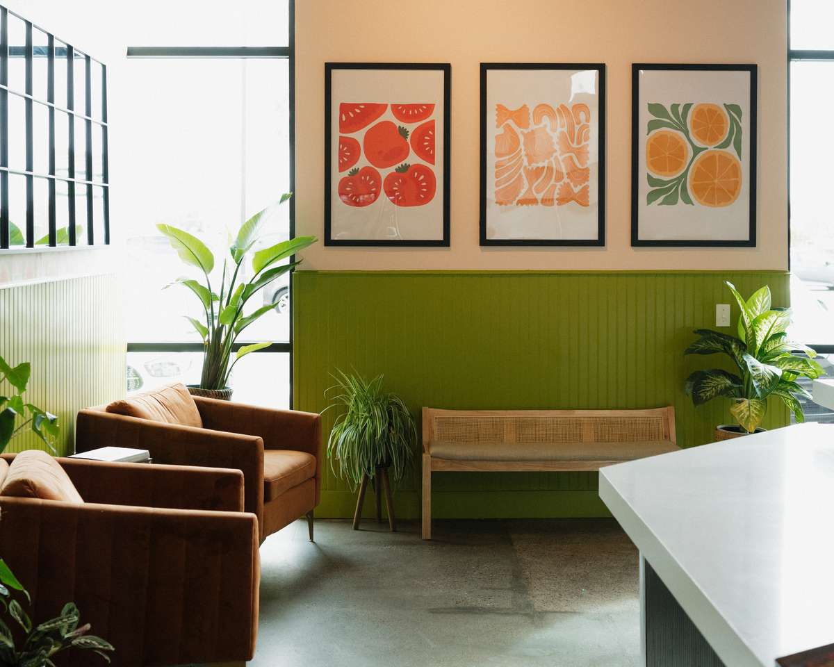 A bright, modern café interior with green and white walls, featuring plants, framed fruit artwork, and a cozy seating area with chairs and a bench.