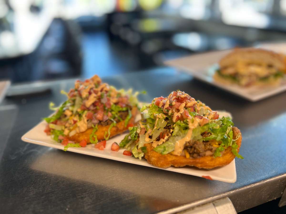 A hand stretched frybread topped with ground beef, lettuce, tomato pico, and cilantro lime crema.