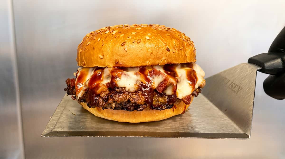 Burger on a metal spatula, with a sesame seed bun, melted cheese, bacon, and barbecue sauce.