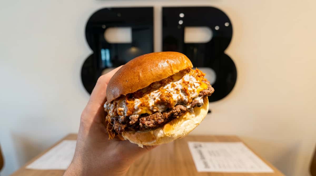 Hand holding a double smash burger with melted cheese, chili, and white sauce on a brioche bun, against a blurry dark background.