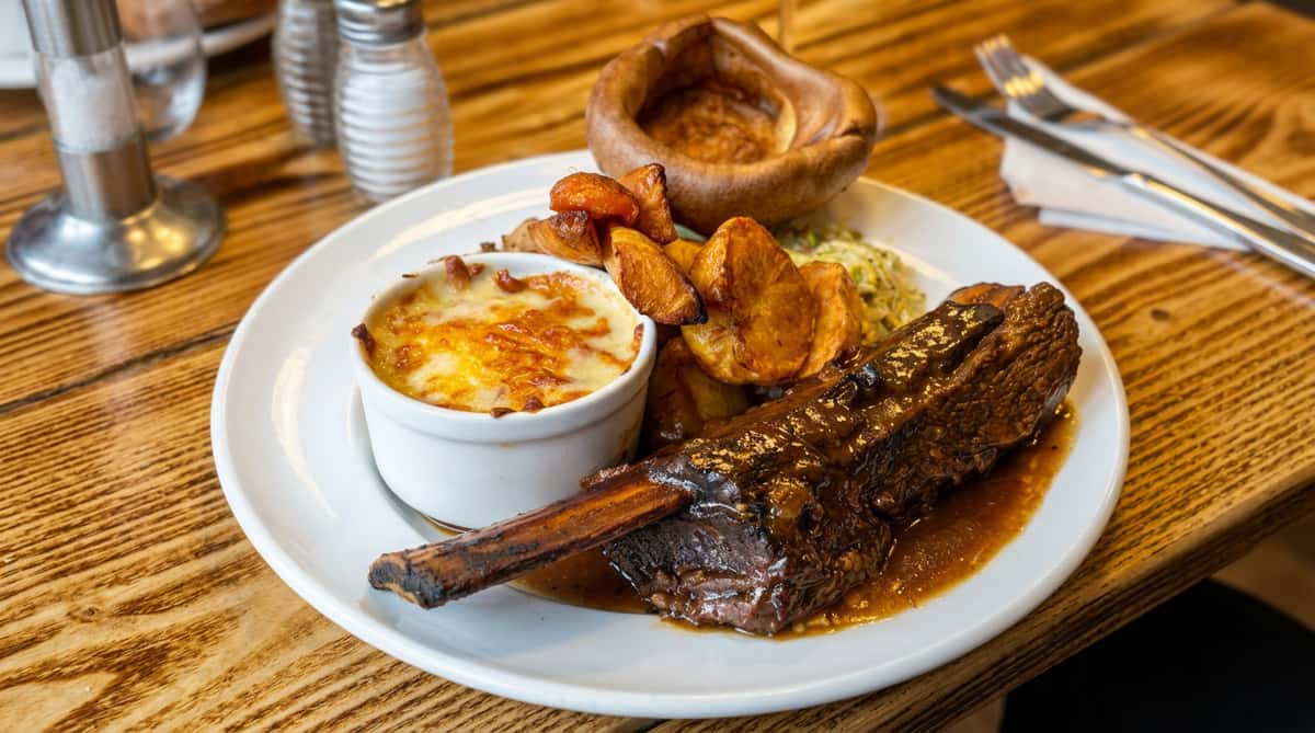 Hearty steak rib with gravy, roasted potato wedges, macaroni and cheese gratin, and a large Yorkshire pudding on a white plate.