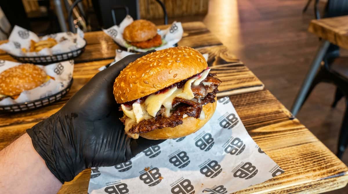 Hand in a black glove holding a beef burger with melted cheese and bacon, a chicken burger in the background.