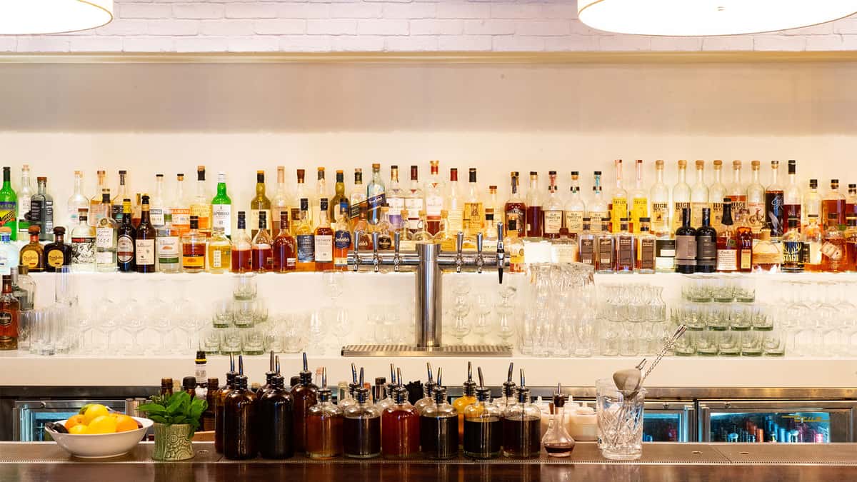 Bright restaurant bar displaying rows of liquor bottles, glassware, and countertop seating.