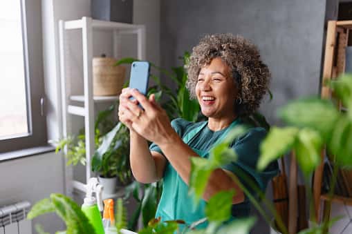 A cheerful woman with curly hair smiles while holding up a smartphone, appearing to take a selfie or video call. She is indoors surrounded by green houseplants, creating a bright and lively atmosphere.