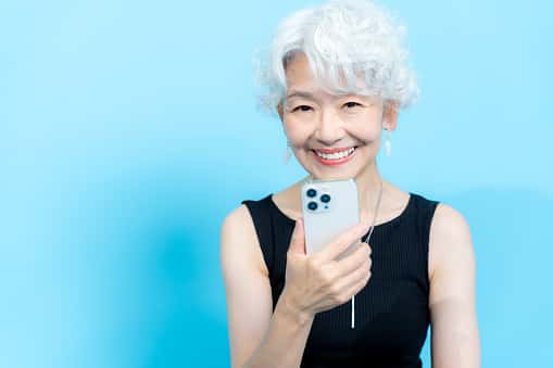 An older woman with short curly white hair smiles warmly while holding a smartphone in her hand. She is wearing a sleeveless black top and dangling earrings, standing against a bright blue background.